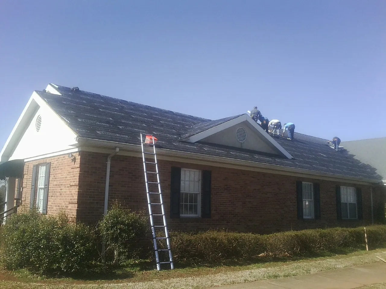 Crew tearing shingles off a brick ranch roof with a ladder set — Emergency Roof Repair work by our crew in Remsen