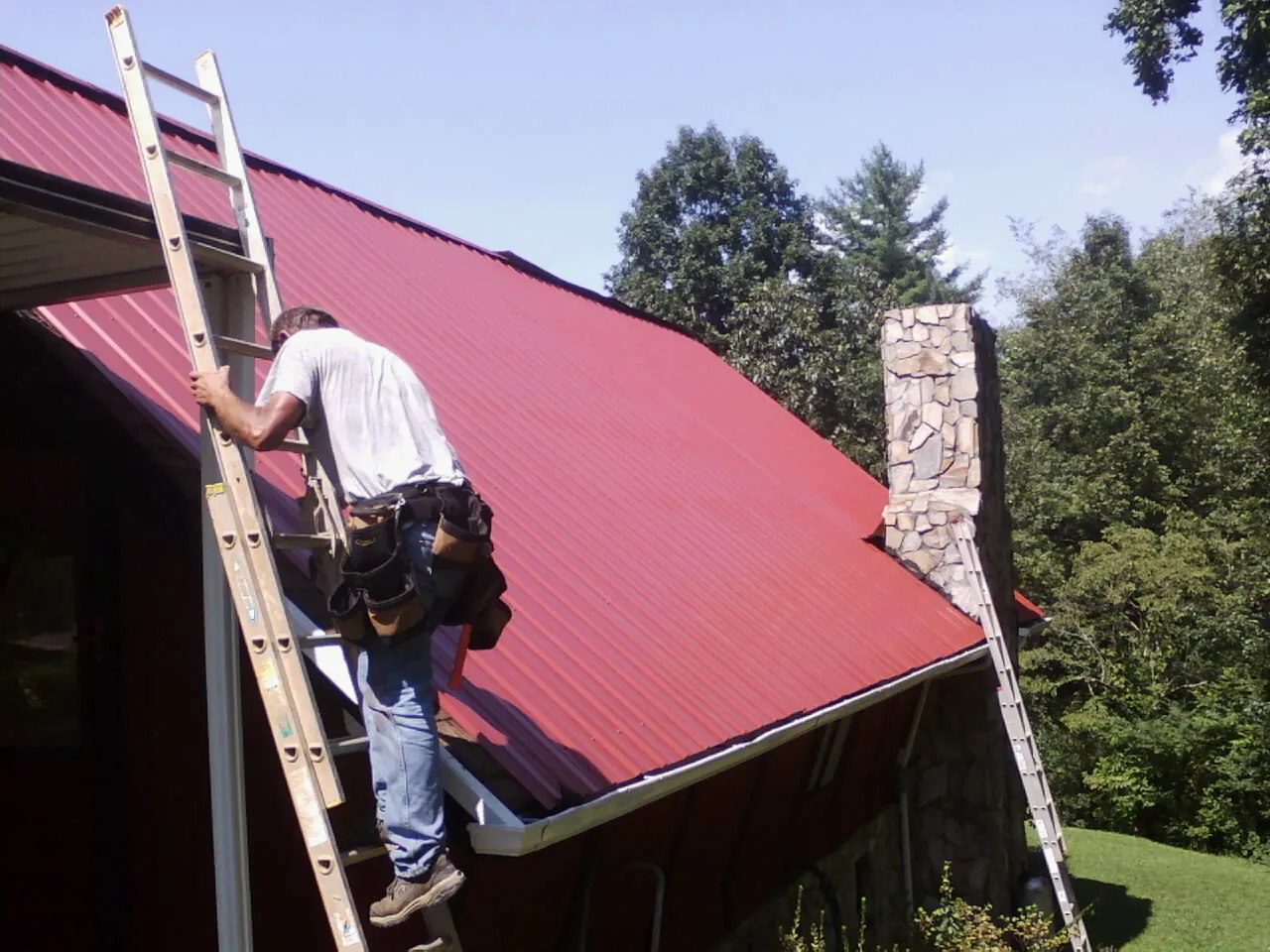 Roofer on a ladder beside a red metal roof and stone chimney — Soffit & Fascia Repair by Roofing Brothers in Remsen